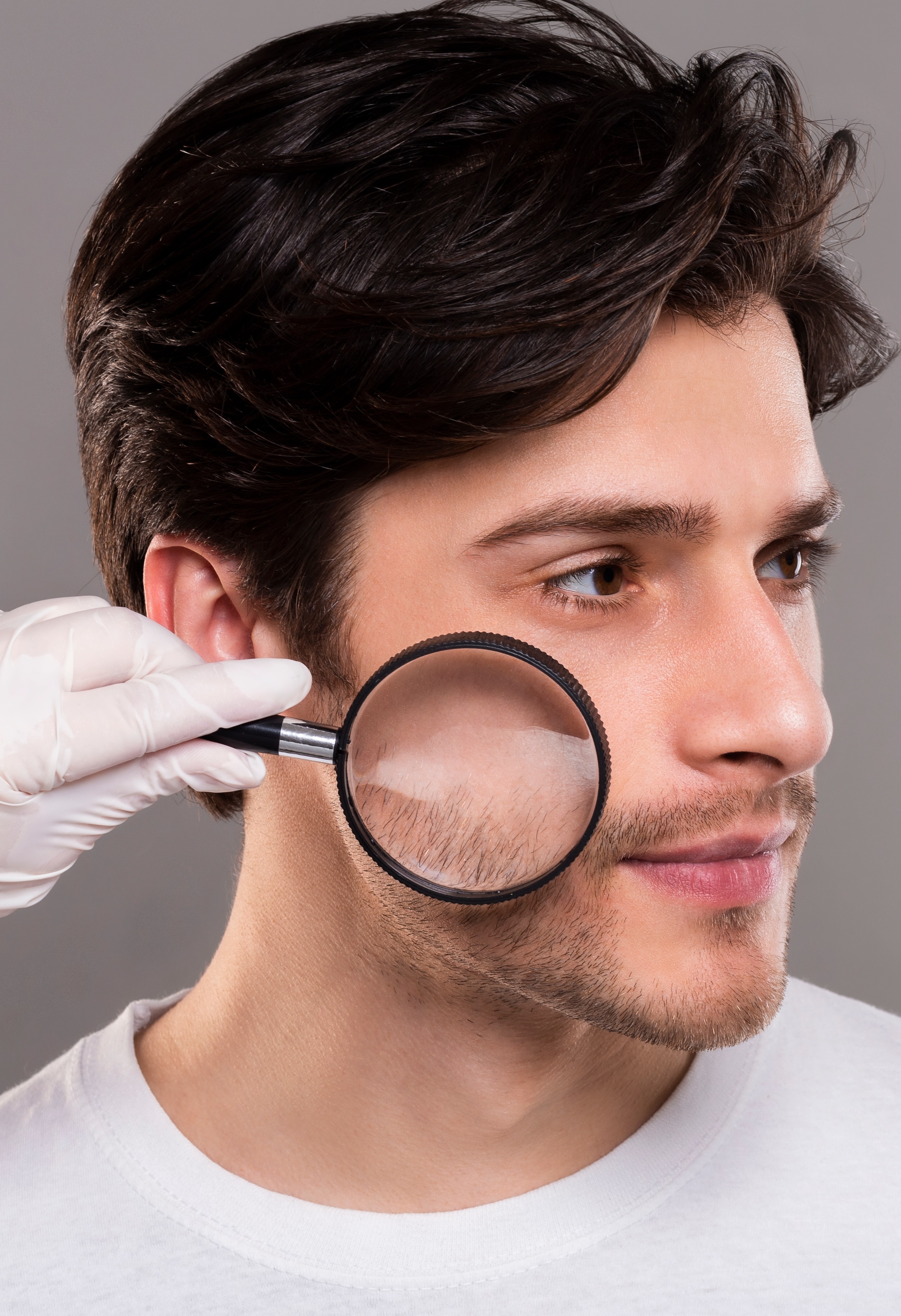 Behandschuhte Arzthand hält Lupe über Gesichtshaut eines jungen Mannes
